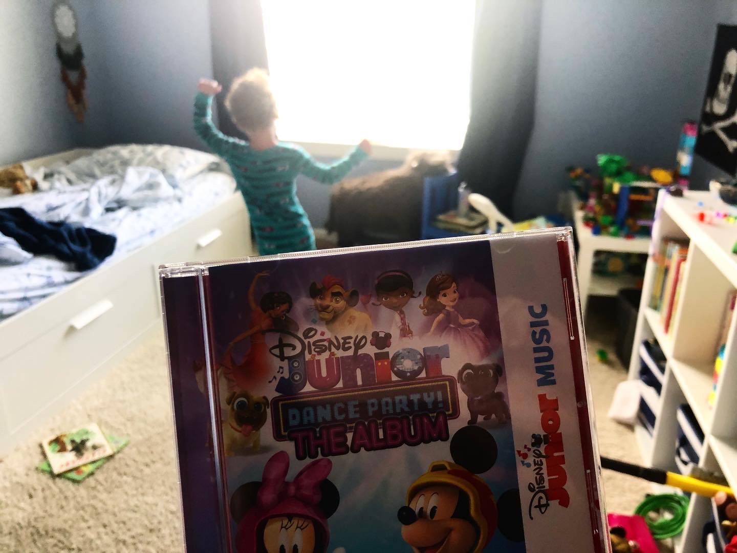 A child is dancing in a messy room with a large open, sunny window. In the foreground of the photo, there is a cd case for Disney Junior songs. Photo by Alyssa Buckley.