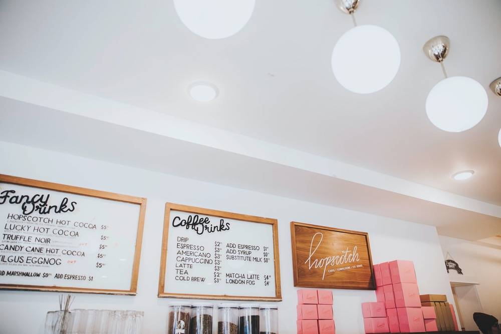 A white wall displaying drink and coffee menus has a shelf of pink paper to-go boxes. There are three circular pendant lights. Photo by Anna Longworth.