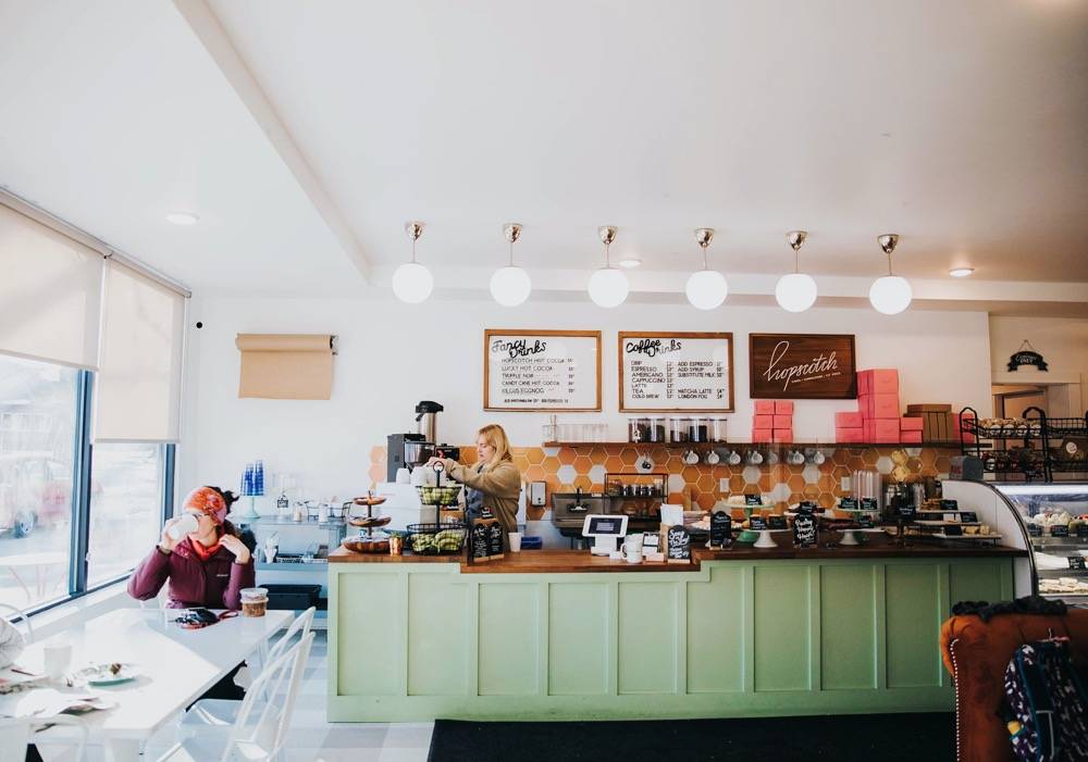 The interior of Hopscotch with patrons sipping coffee at a table by the window and a barista making coffee. The coffee bar is a light green with a glass separator that displays pastries. Photo by Anna Longworth.