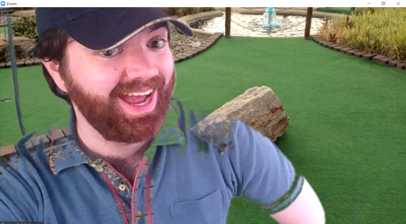 A screenshot of the author in a black baseball cap and blue collared with the mini golf green in the background. Image by Tom Ackerman. shirt