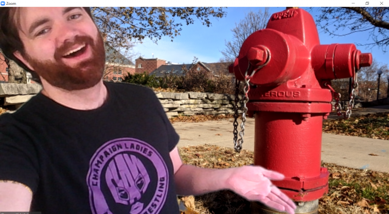 A screenshot of the author in a black t-shirt with a purple Champaign Ladies Amateur Wrestling logo. He is laughing and gesturing towards the fire hydrant. Image by Tom Ackerman.