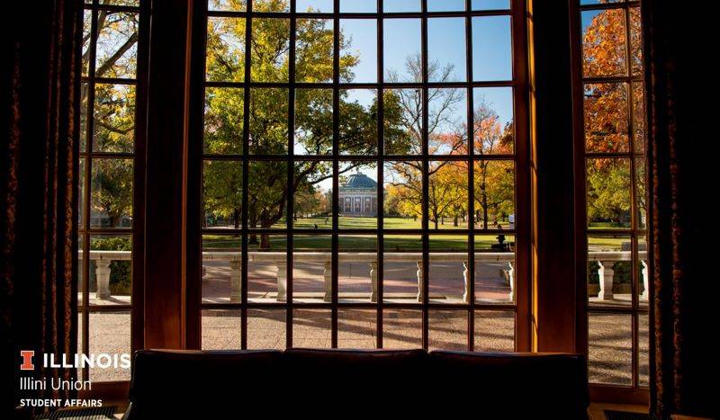 A view through a large window with a grid pattern of window panes. Outside there are leafy trees lining the quad. In the distance is Foellinger Auditorium, a red brick building with white columns and a black dome. Photo from Illini Union Facebook page. 