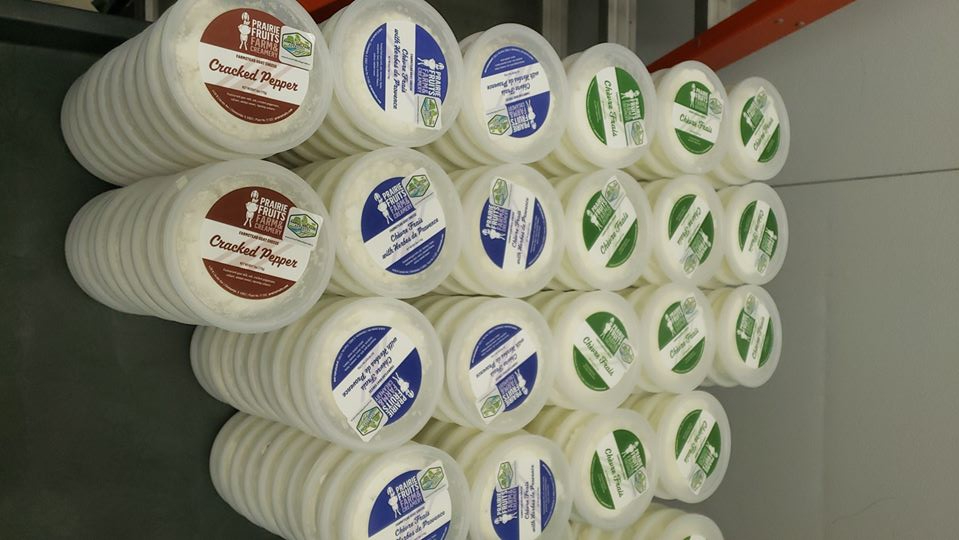 Stacked plastic circular containers of Chevre cheese are lined up on a black table. There are three flavors of different colors: red, blue, and green labels on clear plastic containers containing white cheese. Photo from Prairie Fruit Farms Facebook page.