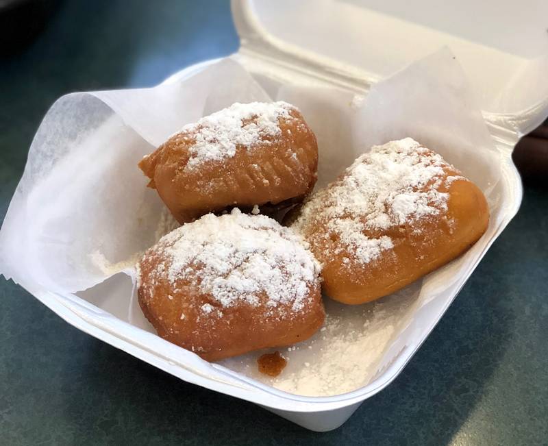 Three beignets topped with a generous amount of powderered sugar sit in a parchment paper lined white styrofoam square container. Photo by Jessica Hammie.