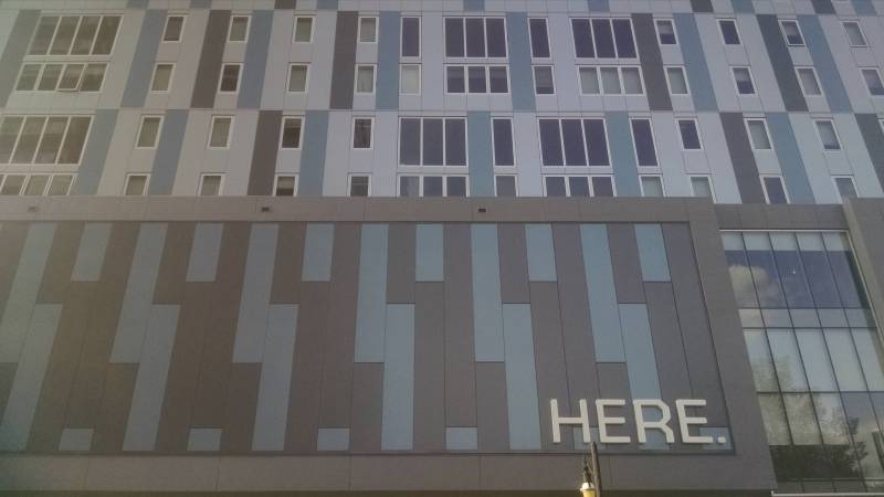 The facade of an apartment building; it's comprised of grayish blue, light gray, and dark gray vertical rectangles, interspersed with windows. In the bottom right corner is the word 