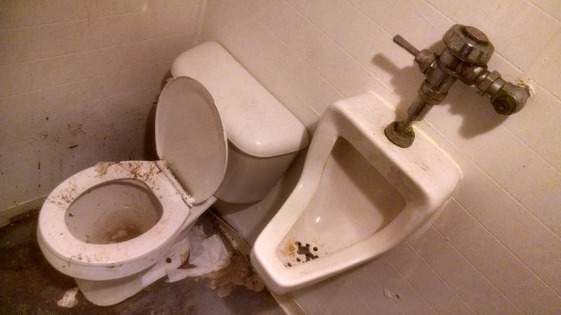 A toilet and urinal covered in brown splatters. The walls are white tile. Photo by Tom Ackerman. 