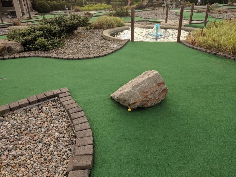 A miniature golf green with a brick border. There is a yellow ball resting against a gray boulder in the middle of the green. Photo by Tom Ackerman.