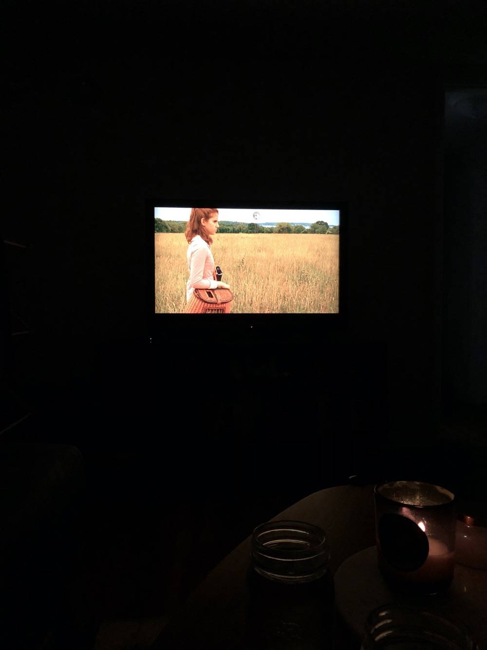 IMAGE: Photo of a distant television screen, featuring a girl standing in a field. There's a candle lit in the foreground of the photo, in a dark room. Photo by Patrick Singer.