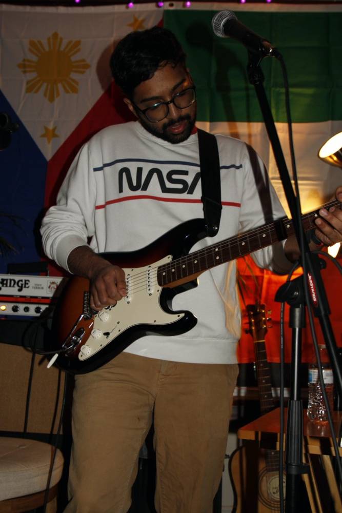 IMAGE: Aditya Kashyap playing guitar, wearing a white long sleeve shirt with NASA's logo on the front, wearing khaki pants. Photo by Matthew Waldinger. 
