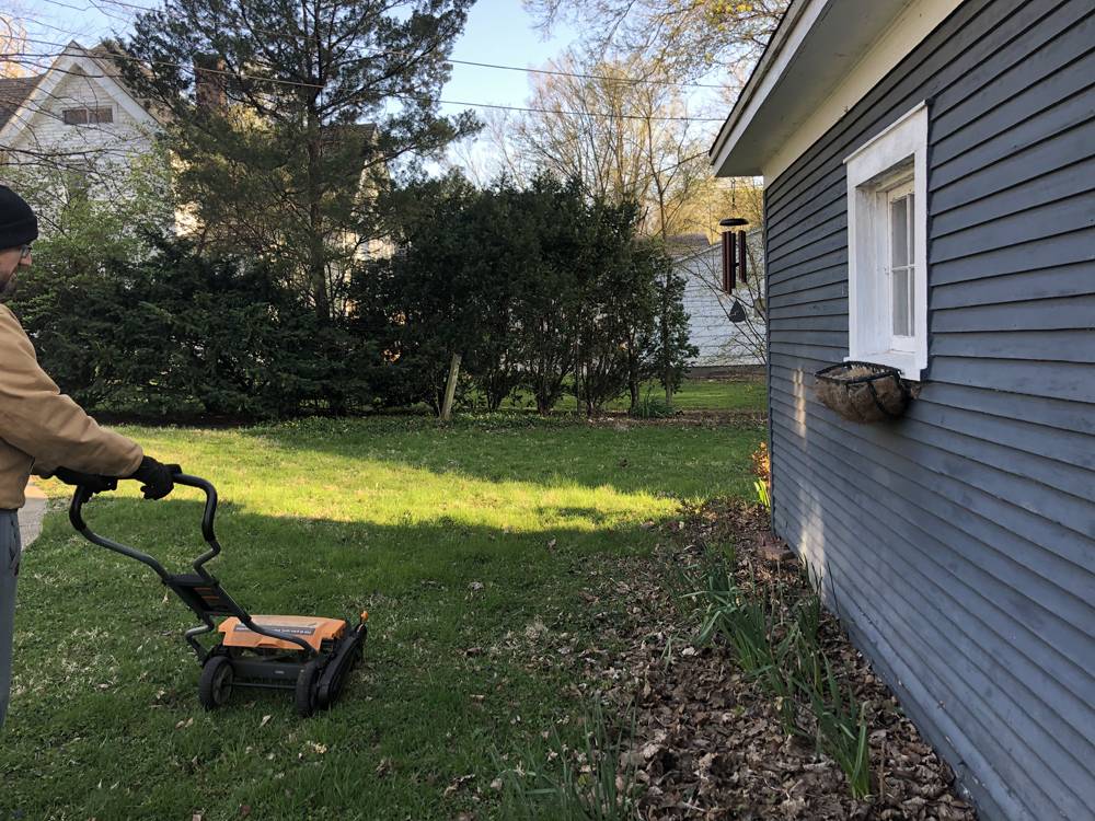 A man pushes a small lawn mower over green grass of a yard. There is a dark gray building to the right of the image. Photo by Carly McCrory-McKay. 