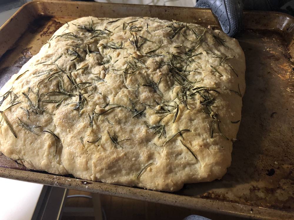 Baked focaccia bread on a sheet pan. A hand with a silicone glove holds the pan. Photo by Carly McCrory-McKay. 