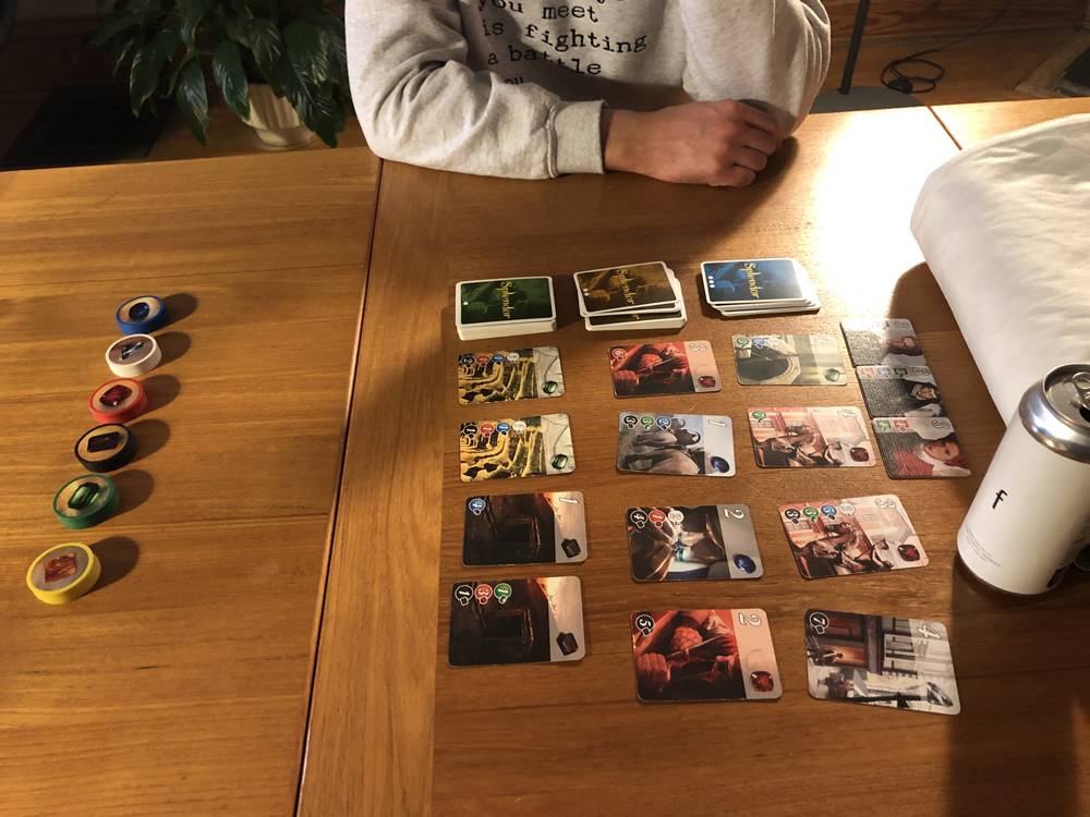 The card game Splendor is laid out in a light colored wood table. Across the table is the arm of a man in a light gray sweatshirt. Photo by Carly McCrory-McKay. 
