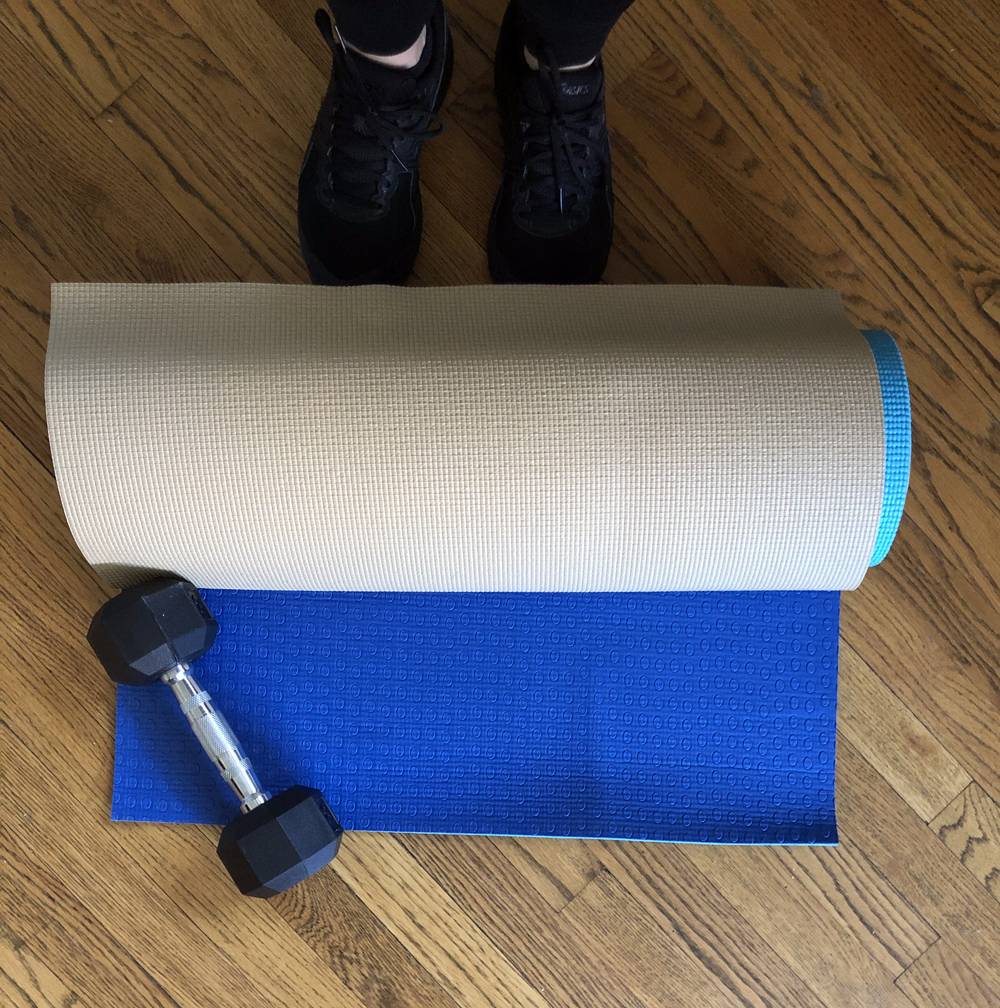Two exercise mats, one blue and one beige, are rolled up and sitting on a wood floor. There is a dumbbell with a chrome handle and black weights sitting on it. A person's feet in black tennis shoes are standing next to the mat. Photo by Jessica Hammie.