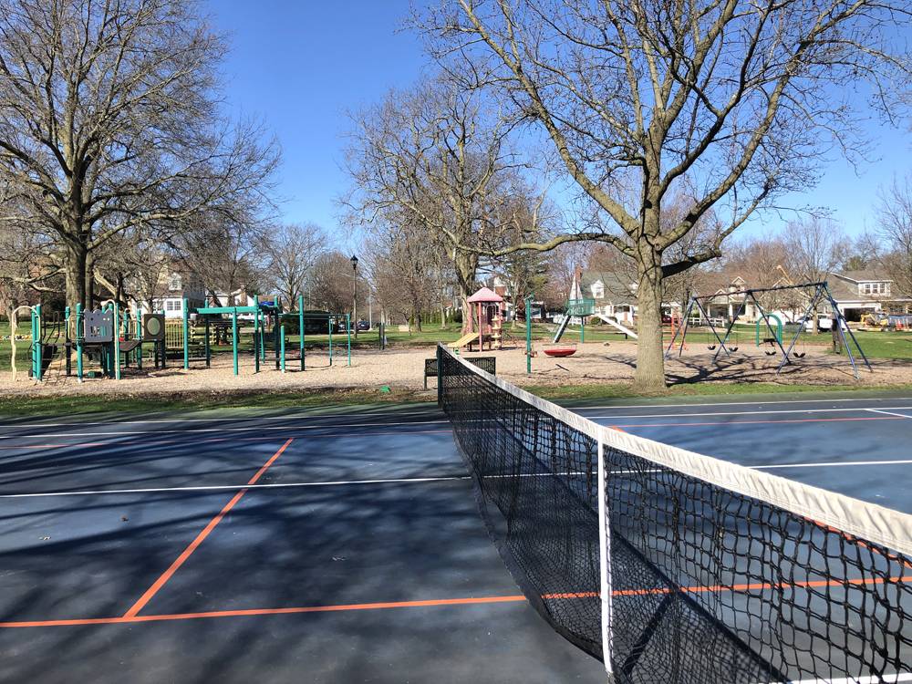 Clark Park tennis court and view of playground. In the foreground, a blue tennis court with white and orange pickleball lines are visible. The net is just off center to the right. The playground and green space is in the backgroudn beyond the tennis court. Photo by Jessica Hammie. 