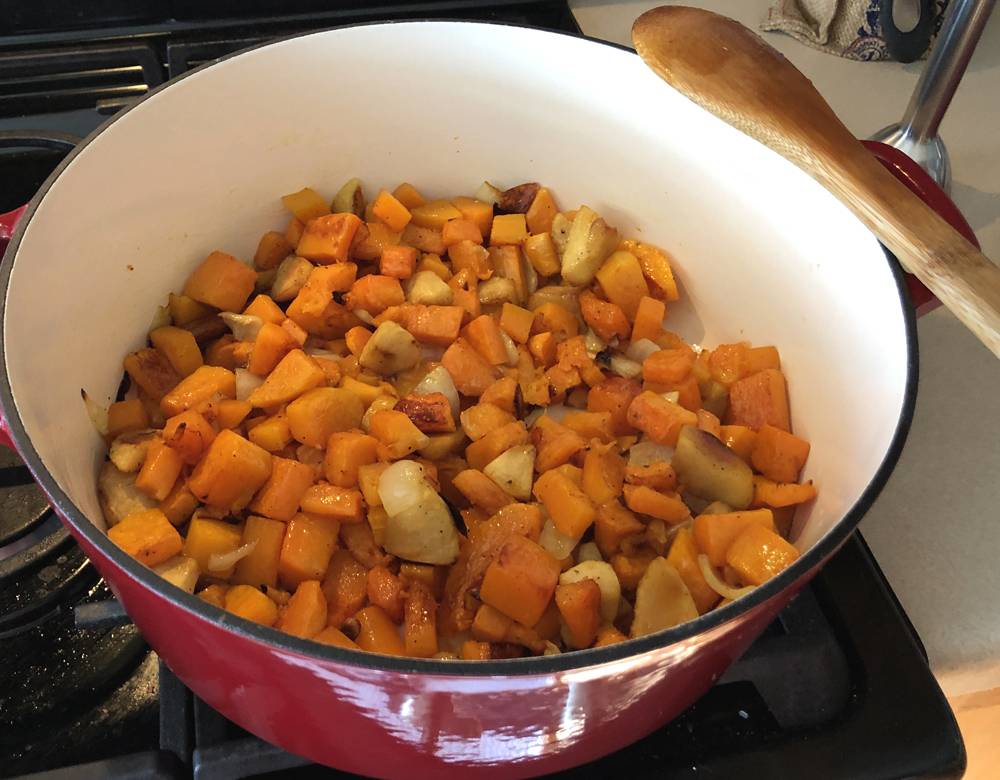 A large red Dutch oven on a gas stovetop. Inside the pot are cubed pieces of butternut squash, onions, and apples. A wood spoon rests across the right side of the pot. Photo by Jessica Hammie. 