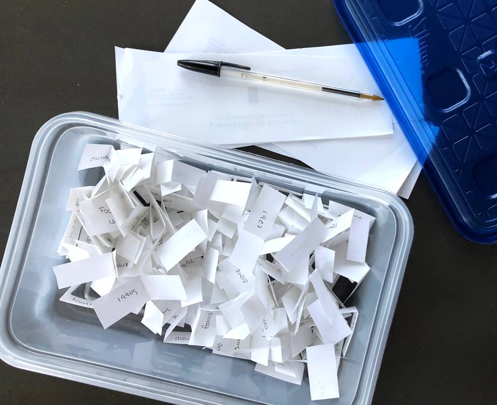 A rectangular plastic container without a lid on it sits on a gray table. Inside the container are slips of paper with words and numbers on them. Also on the table are the lid to the container, slips of paper, and a ball point pen. Photo by Jessica Hammie.