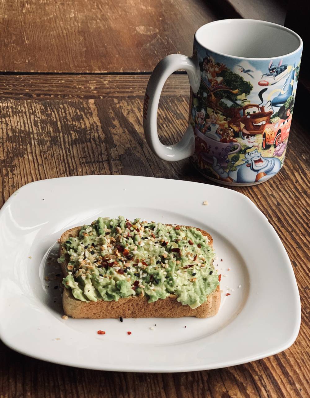 A slice of toast on a white plate. The toast is dressed with smashed avocado and seasoning. There is a Disney-themed mug behind the plate. Everything sits on a wood table. Photo by Stephanie Stuart.