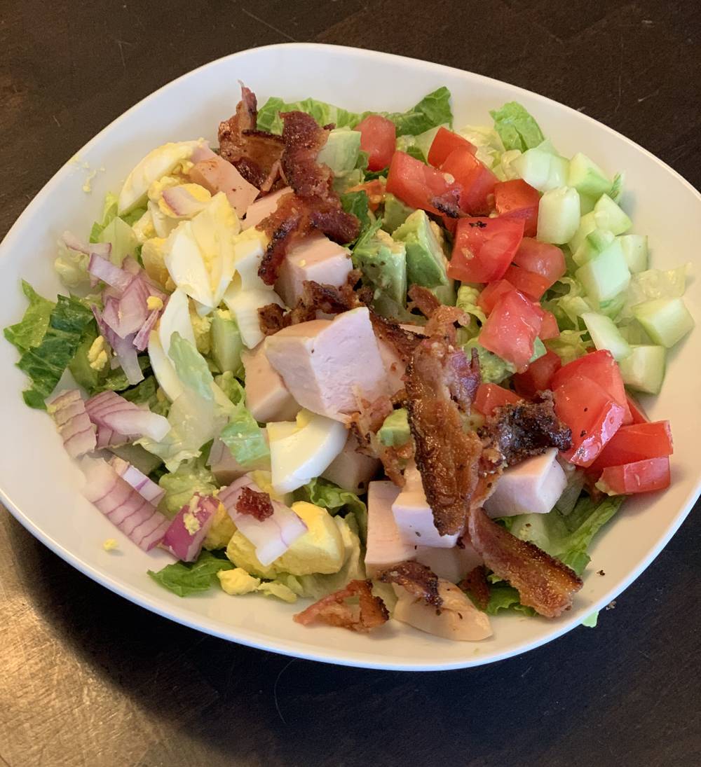 A homemade Cobb salad in a white bowl. The salad contains shredded greens, red onion, hard boiled eggs, large chunks of chicken, bacon, avocado, tomato, and cucumber. Photo by Stephanie Stuart.
