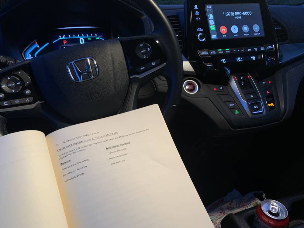 A view from the driver's seat of a car. The driver (not pictured) is holding a book open. It is night, and the interior car lights are on. There is a phone number on the info screen. The Honda logo is visible on the stearing wheel. Photo by Stephanie Stuart.