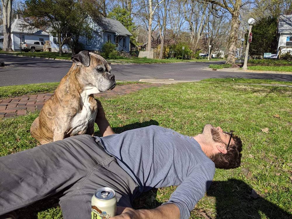 A man lays in the grass. Next to him is an old dog who looks out over the yard. The man has one hand on the dog, and holds a beer in the other.  Photo by Rey Dalitto. 