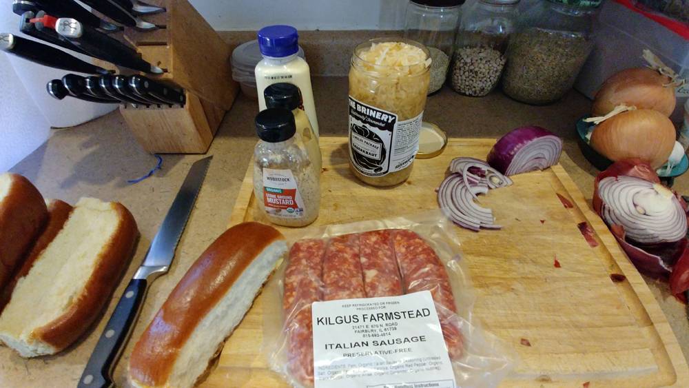 A cutting board in a kitchen holds a package of Kilgus Farmstead Italian sausage, some bread, kraut, and several other condiments. There is a large bread knife on the counter, and a knife block behind it. Photo by Taidghin O'Brien.