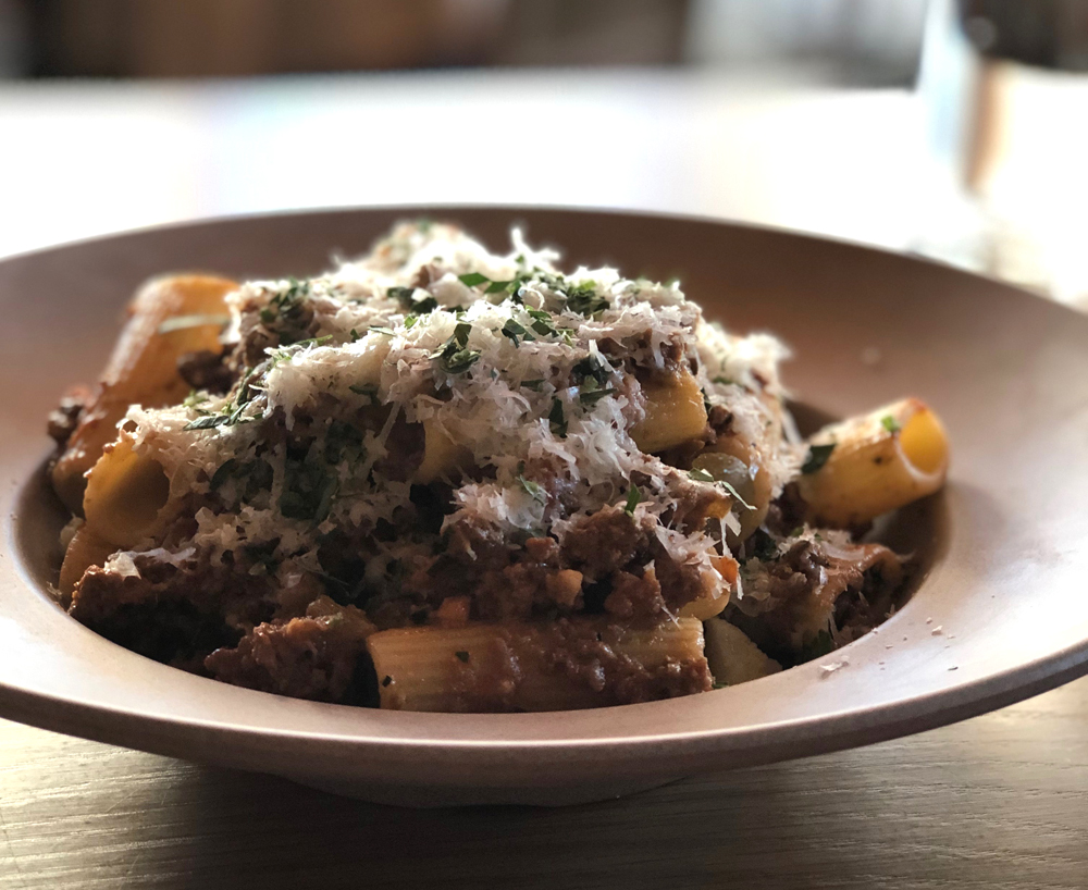 A portion of rigatoni pasta tossed in a bolognese sauce with grated parmesan and chopped herbs on top sits in a gray circular bowl on a wooden table. Photo by Jessica Hammie.