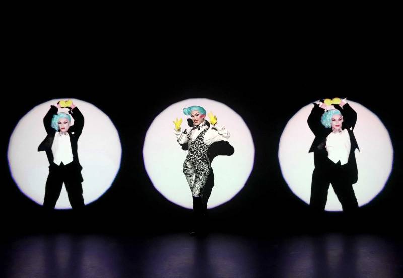 Drag queen Sasha Velour is represented three times in three different spotlights. The Sasha in the center is wearing a white blouse and leopard print jumpsuit. The Sashas on the left and right are wearing black tuxedos with white vests. She has a bright blue wig. Photo by Jeff Eason.