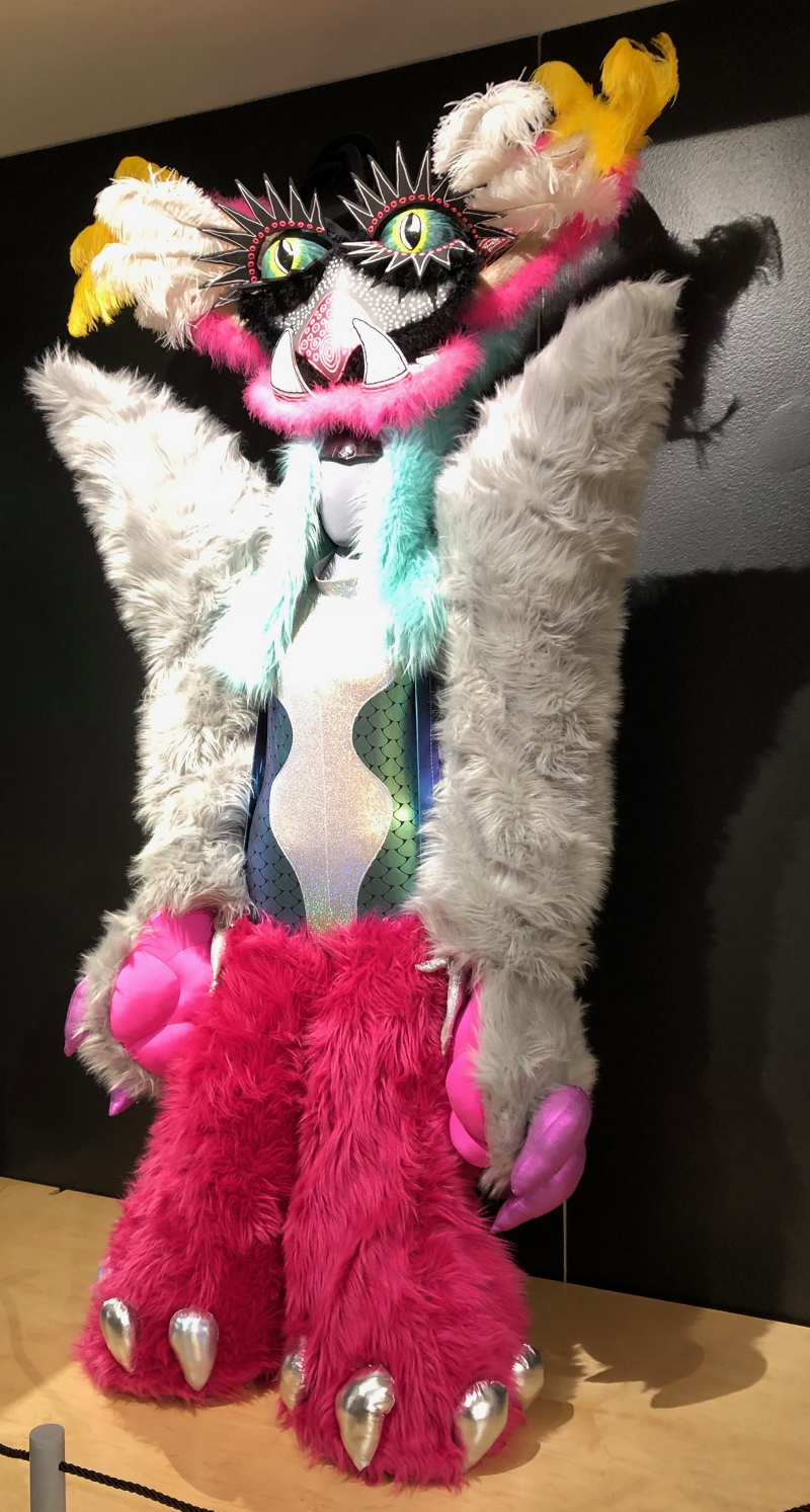 A large fanciful costume that is covered in fur. There is a headpiece with fangs and large green and yellow eyes with exaggerated black eyelashes. White and yellow feathers extend from either side. The body has white furry arms and pink furry legs. Photo by Cope Cumpston. 