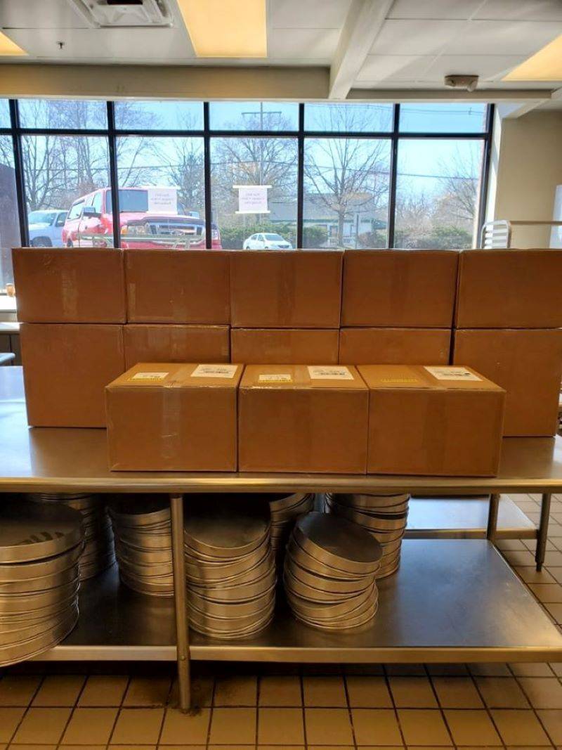 A stack of cardboard boxes sit on a counter. They are taped and ready for shipping. Underneath the counter there are stacks of metal pizza pans. Photo provided by Papa Del's. 