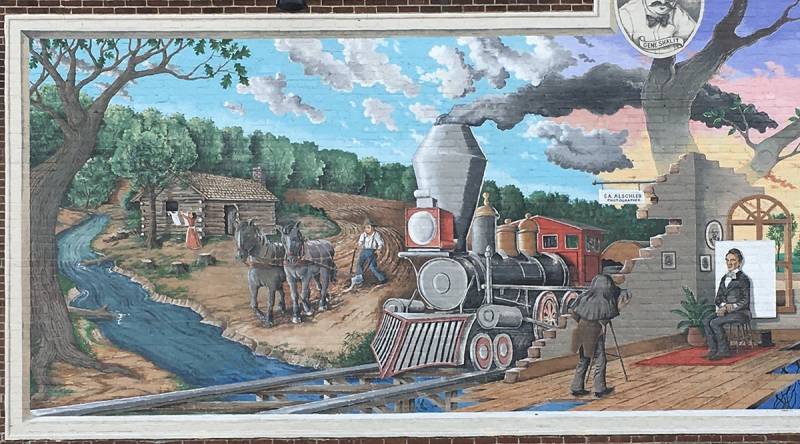 The east section of the mural pictured is about 12' x 5', depicting a log cabin by a winding stream with a forest in the background below white clouds on a blue sky.  Tompins walks behind a plow drawn by two gray horses across a furrowed field.  His wife hangs clothes on a line in the background.  To his left, in a shift of scenes, a locomotive comes into 
