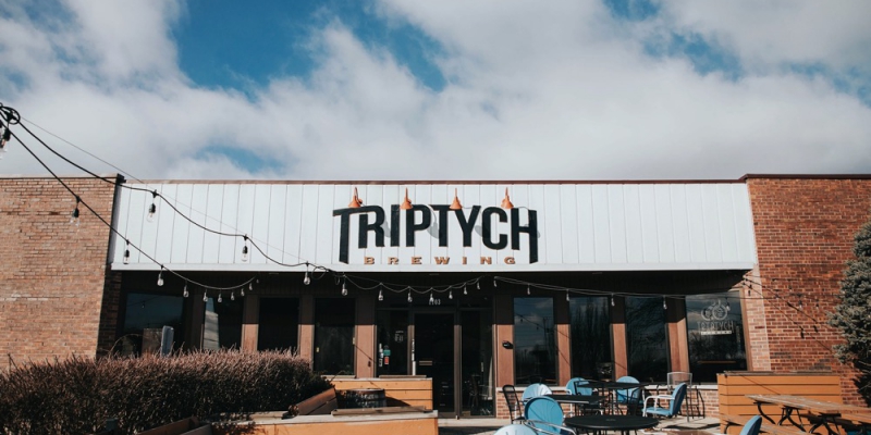 An outdoor shot of the Triptych Brewery with the placard centered on a white vertical shiplap storefront. There are string lights, bushes, and outdoor tables also viewable. Photo by Anna Longworth.