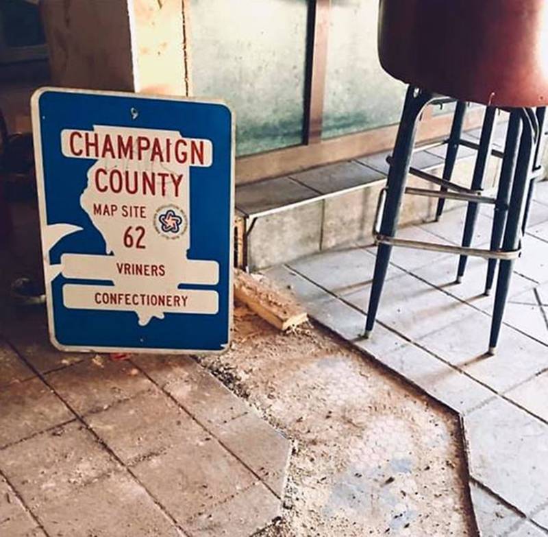 Commemorative sign for Vrinerâ€™s Confectionary, 24â€ tall x 18â€ wide, sitting on a tile floor, leaning against the wall of the old building in downtown Champaign. Red lettering on a white silhouette of Illinois set on a blue background reads, â€œChampaign County Map Site 62, Vrinerâ€™s Confectionary. Image from Champaign County History Museum. 