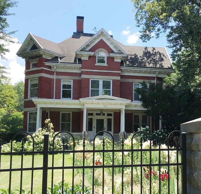 A large, red brick building; two main floors with a gabled third floor and window dormers. A white frame porch runs across the front of the house, which sets on the south side of the street on a sunny summer day, fronted by a beautifully landscaped lawn. The houseâ€™s style is eclectic in nature, typical of Midwestern architecture in the early 20th Century. Photo by Rick Williams.