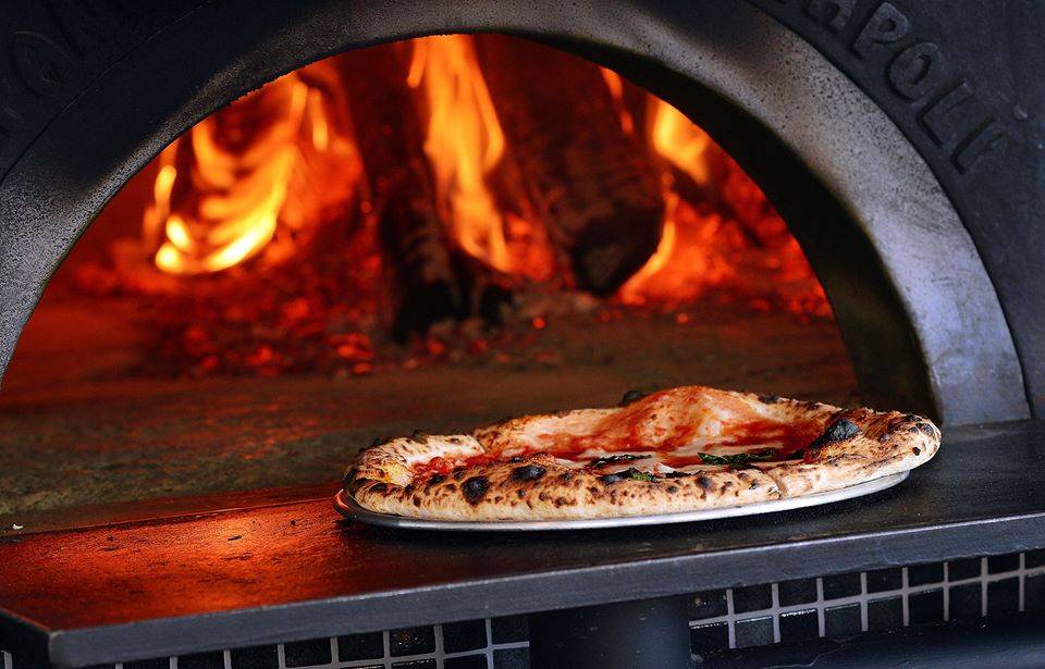 A cooked pizza sits on a circle, metal pan outside of a huge, black firey pizza oven. Photo by Pizzeria Antica.