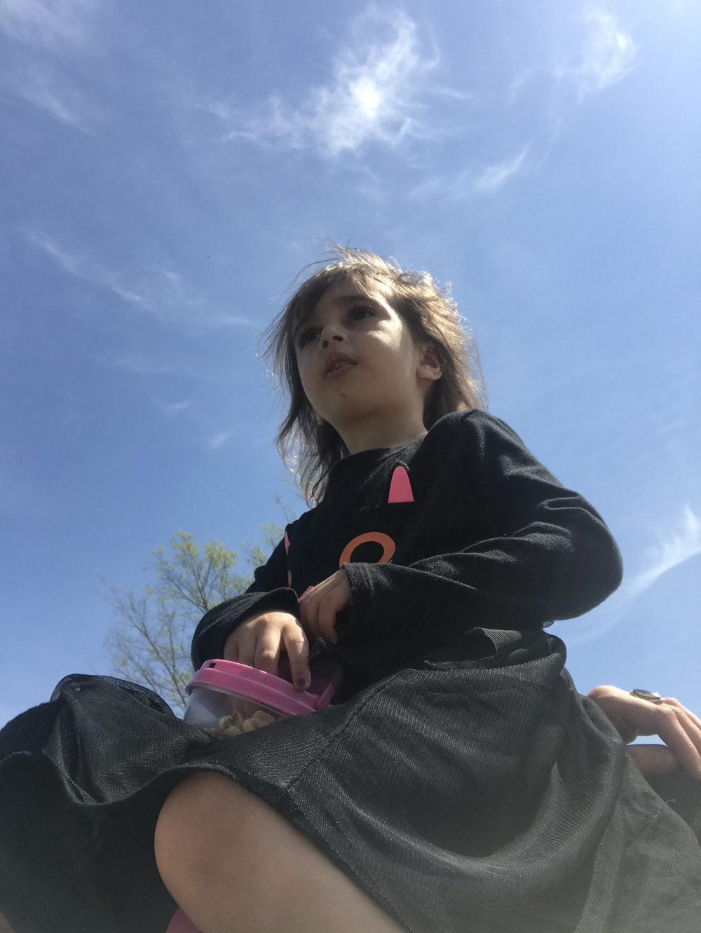 A toddler girl wearing a black cat shirt and a black skirt is photographed from below. She is sitting down and looking off into the distance. In her hands is a container full of snacks. Photo by Darya Shahgheibi.