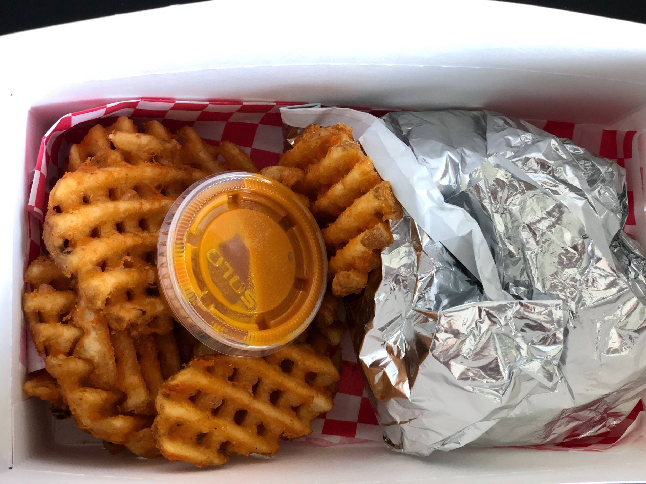 A togo order from Watson's: waffle fries with a small cup of honey mustard sits in a white paper box next to a fried chicken sandwich wrapped in a metallic wrapper. Photo by Alyssa Buckley.