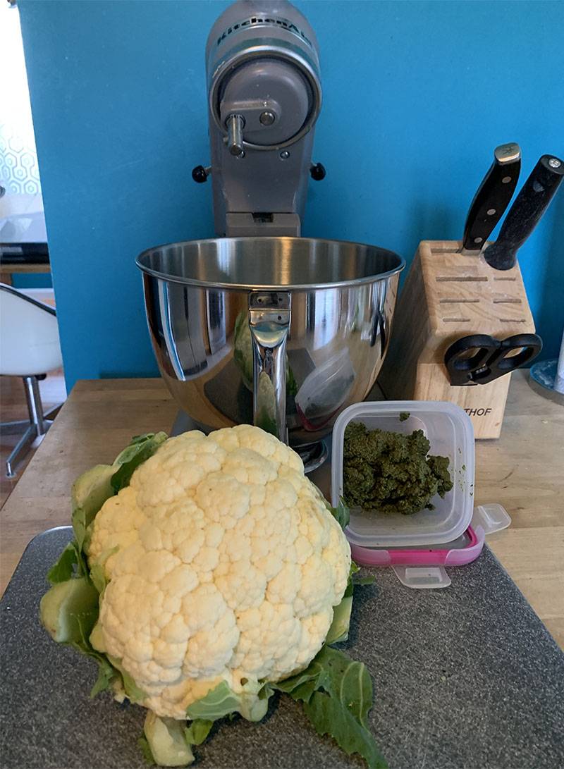 On a kitchen counter is a stand mixer, a cutting board with a head of cauliflower, and a container of pesto. The wall in the background is bright blue. Photo by Debra Domal. 