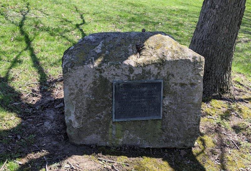 A large stone marker sits in the shadow of a tree. It has a metal plaque in the center. Photo by Katriena Knights. 
