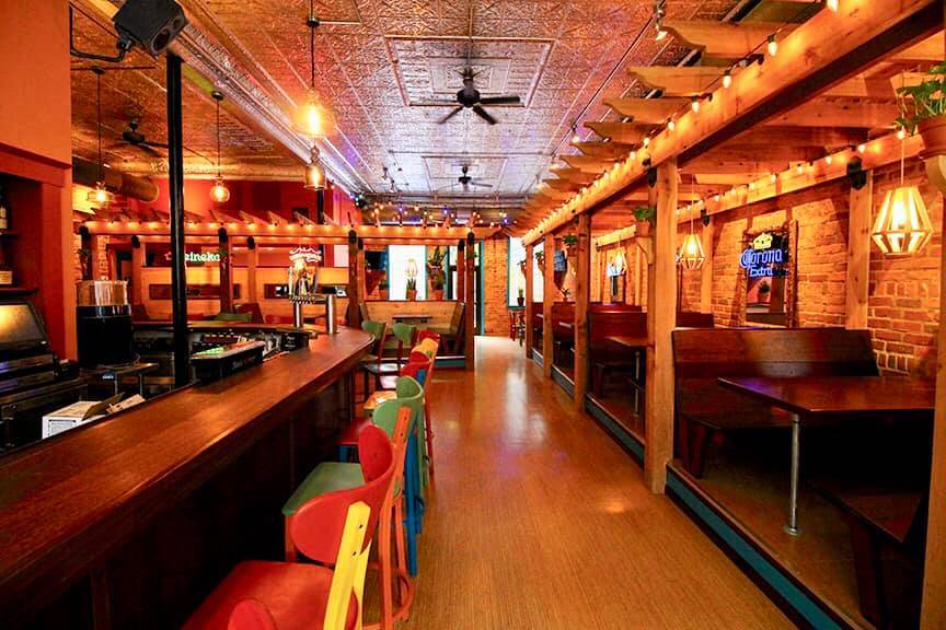 A photo of the interior of Cowboy Monkey. The differently colored barstools are empty at the bar on the left, and booths are empty on the right. Photo from Cowboy Monkey's Facebook page.