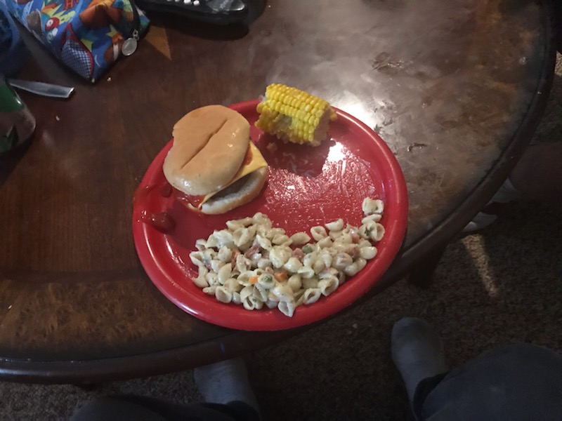 A red paper plate sits on a round table. On the plate is a cheeseburger, a small section of an ear of corn, and pasta salad. Photo by Gillian Dabrowski. 