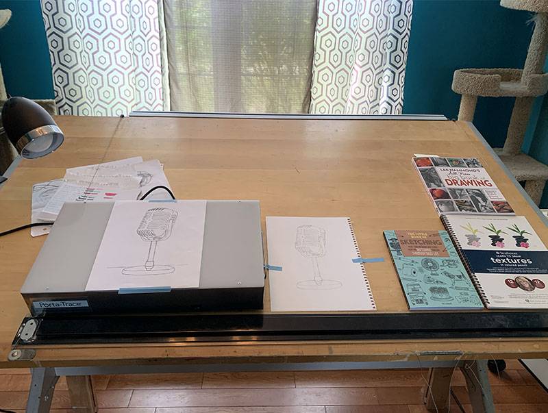 A light wood colored crafting table is in front of a window with curtains. The walls are bright blue. On the desk is a tracing light box, two drawings of a microphone, and some books. Photo by Debra Domal.