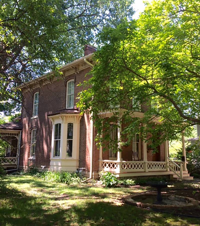 Two-story brick with bracketed roof overhanging 2 feet.  Large frame bay window, painted soft yellow, juts out on left side, first floor.  Open frame porch extends across the front and wraps around the right side.  Surrounded by green plants, shrubs, trees. Photo by Rick D. Williams. 