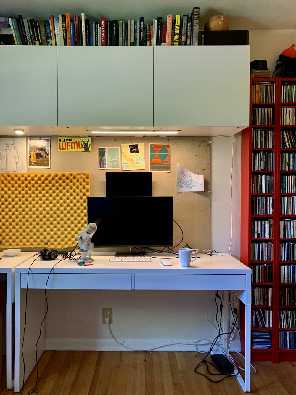 View of a desk in a home office. On the white desk is a microphone, a computer monitor, keyboard, and mouse. There are books on a shelf above the desk, and another bookshelf with books to the right of the desk. On the wall behind the desk is a pin board, with postcards and noise-buffering foam. Photo by Kevin Hamilton. 