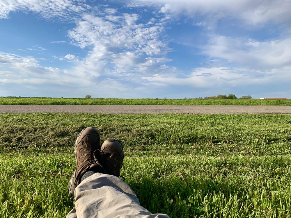 The lower legs and feet of a man are pictured in repose on a green field. Just in front of the person is a paved pathway, horizontal across the picture. Beyond that is more green, and a very blue sky with whispy clouds. Photo by Kevin Hamilton. 