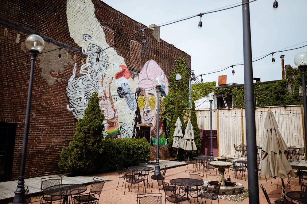 A patio with black tables and chairs. There are string lights hanging above the seating, as well as a few beige umbrellas. Thereâ€™s a mural painted on a brick wall in the background. Photo by Anna Longworth.