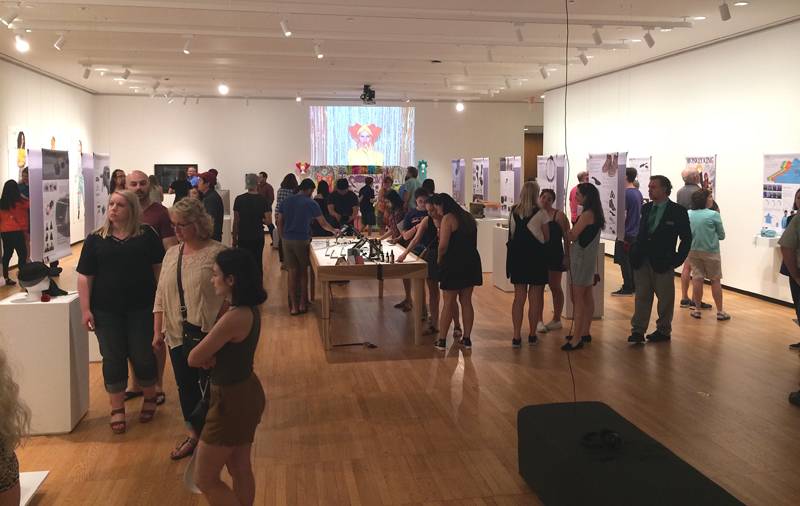 Gallery at Krannert Art Museum during the opening reception of the 2018 School of Art + Design BFA Exhibition. In the large gallery are wall hung artworks and artwork on pedestals. There are a lot of people in the room looking at the various displays. Photo by Jessica Hammie. 