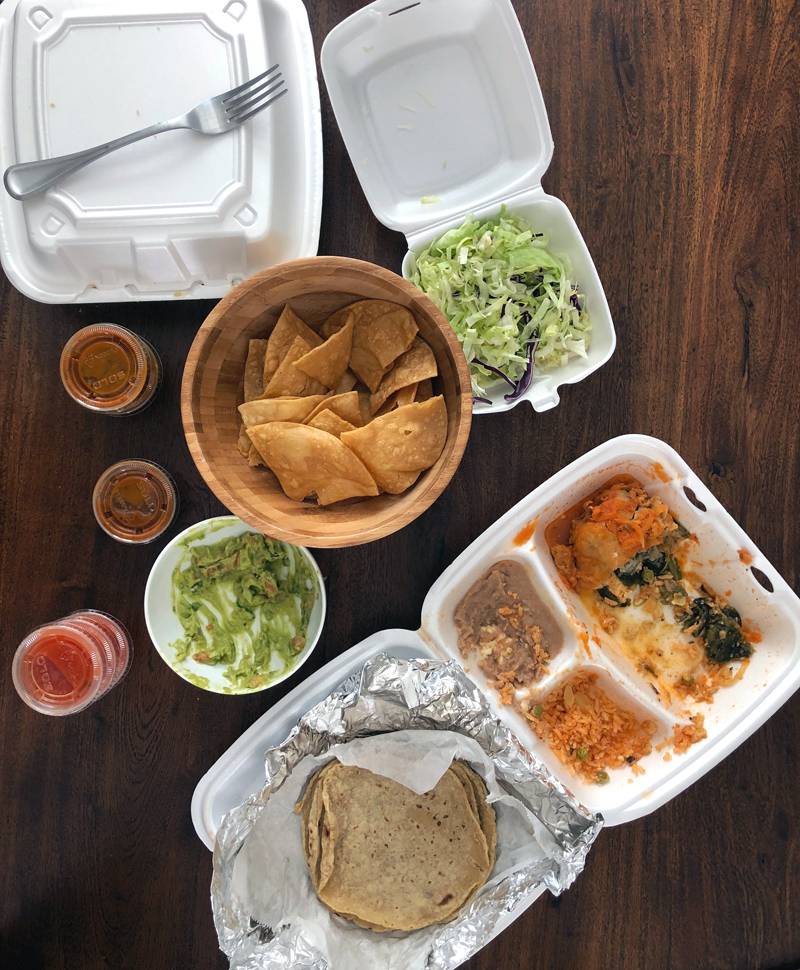 An overhead photo of takeout food: a close styrofoam container has a metal fork on top. There are small cups of sauce with covers on them. A bowl of tortilla chips is in the center. An open styrofoam container of corn taco shells with rice and fillings is mostly eaten. Photo by Jessica Hammie.