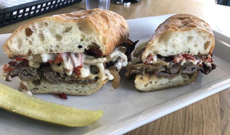 A sandwich on a bageutte is sliced in half and you can see melty white cheese, red peppers, and beef. There is a pickle next to the sandwich on a piece of parchment paper. Photo by Jessica Hammie.