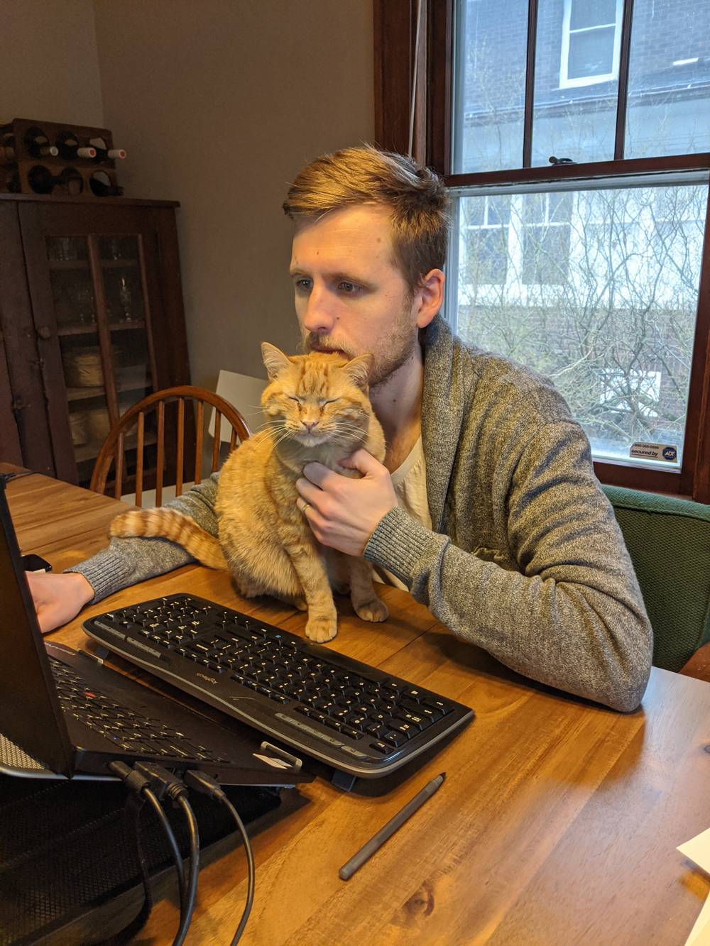 Jonathan Hafer is seated at a wood dining table. On the table is an orange cat. Hafer has his left hand on the cat, and the cat's eyes are closed. The cat looks blissed out. Hafer is working at at computer. Photo courtesy of Jonathan Hafer. 