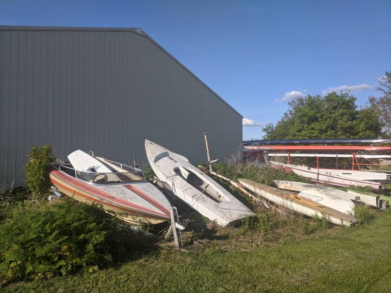 Several damaged boats are lined up in front of a gray warehouse building. There is overgrown brush all around them. Photo by Tom Ackerman.